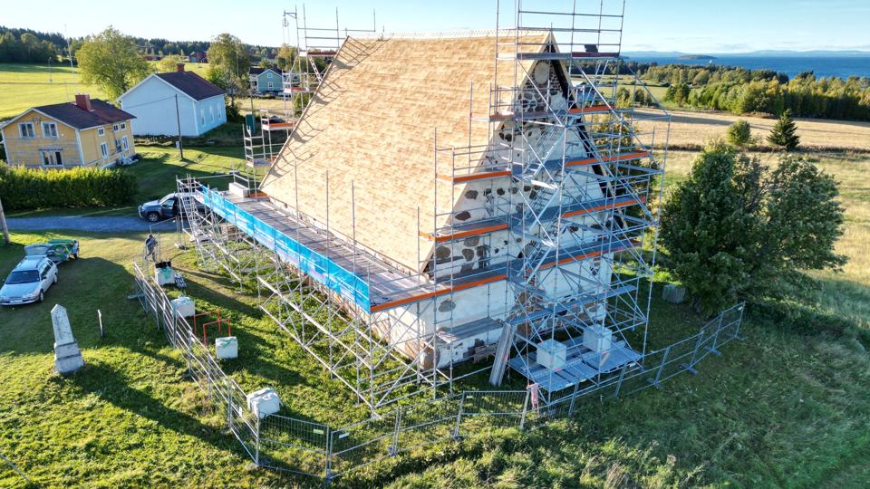 Drönarbild: rött hus med ställning längs taket mitt i ett sommarlandskap