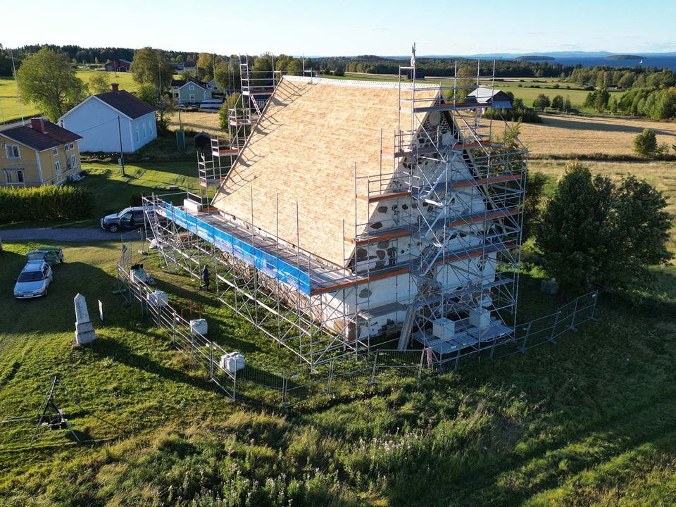Drönarbild över ett hus vars gavel är inbyggd i byggställning, med jämtländskt odlingslandskap runt omkring